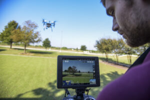 Photo of a researcher viewing a drone’s live video feed on a handheld monitor at Texas A&M’s RELLIS Campus. A drone is seen flying above a grassy field with trees and blue sky in the background.